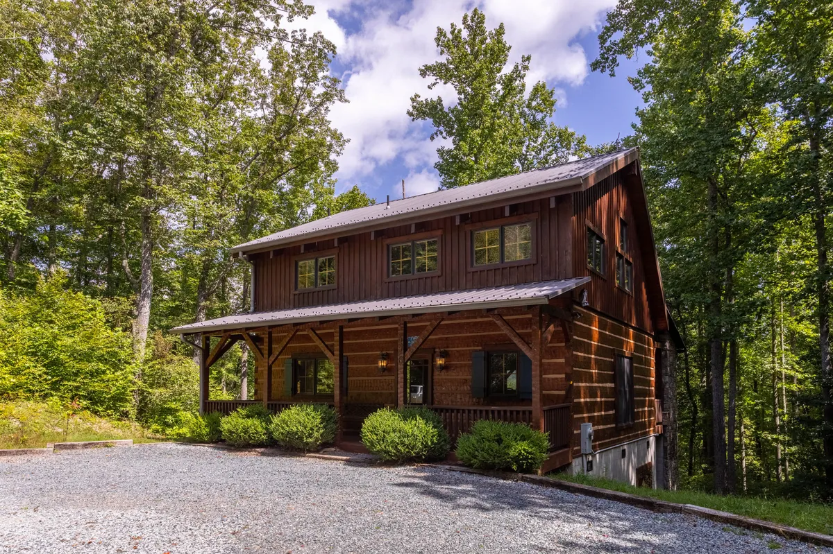 The image showcases a charming rustic wooden cabin nestled among tall trees, with a welcoming front porch and a gravel driveway leading up to the home. The cabin exudes tranquility and blends harmoniously with the natural surroundings, inviting visitors to relax and soak in the peaceful mountain atmosphere.