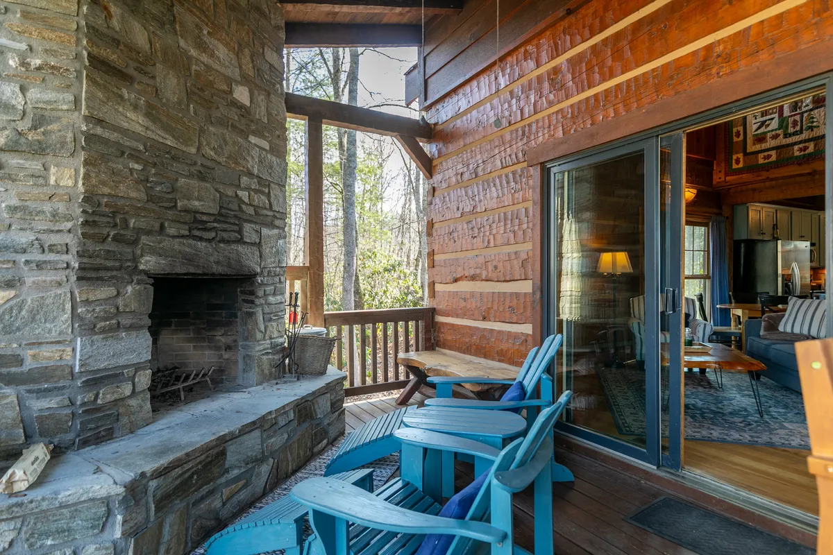 This image presents a deck scene with a striking stone fireplace, blue chairs, a sturdy wooden railing, and vast nature views in the background. The setup encourages leisurely mornings or cozy evenings by the fire.