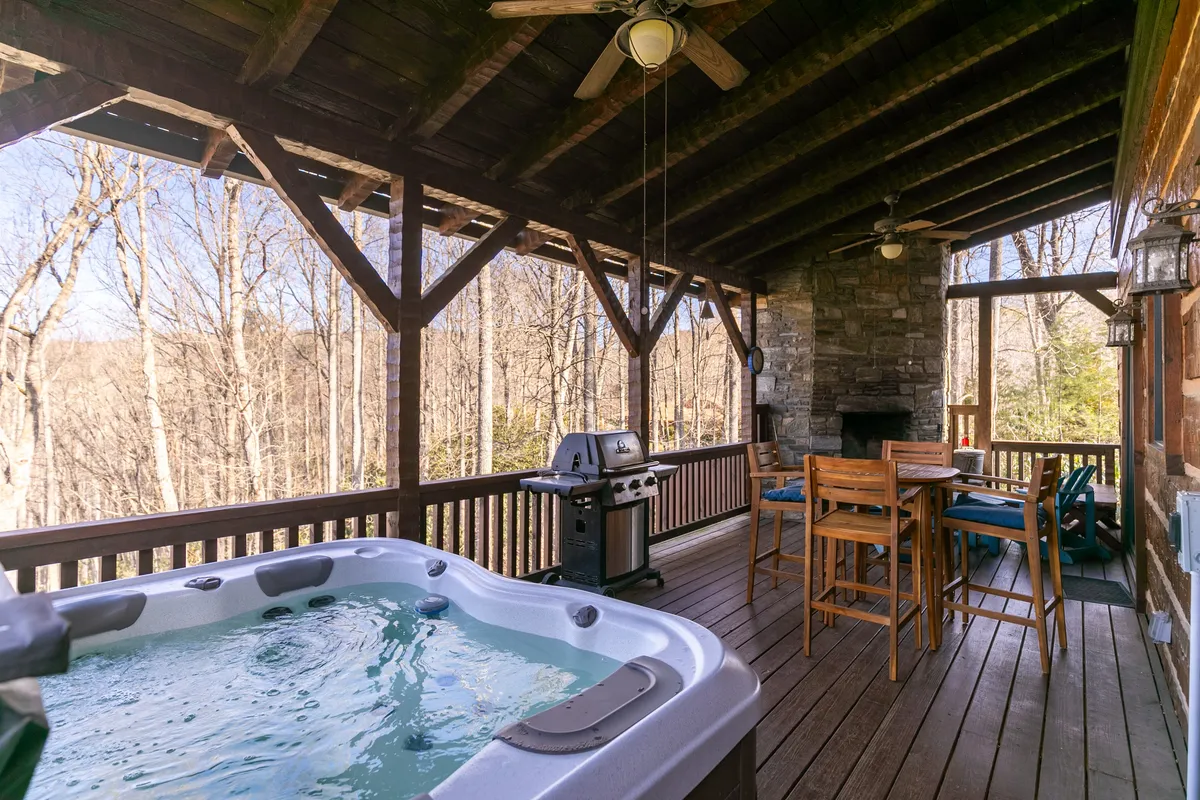 A vibrant outdoor scene showing a hot tub, gas grill, and a dining set arranged on a spacious wooden deck, with lush trees all around. The setup suggests opportunities for both soothing soaks and al fresco meals among the forest backdrop.
