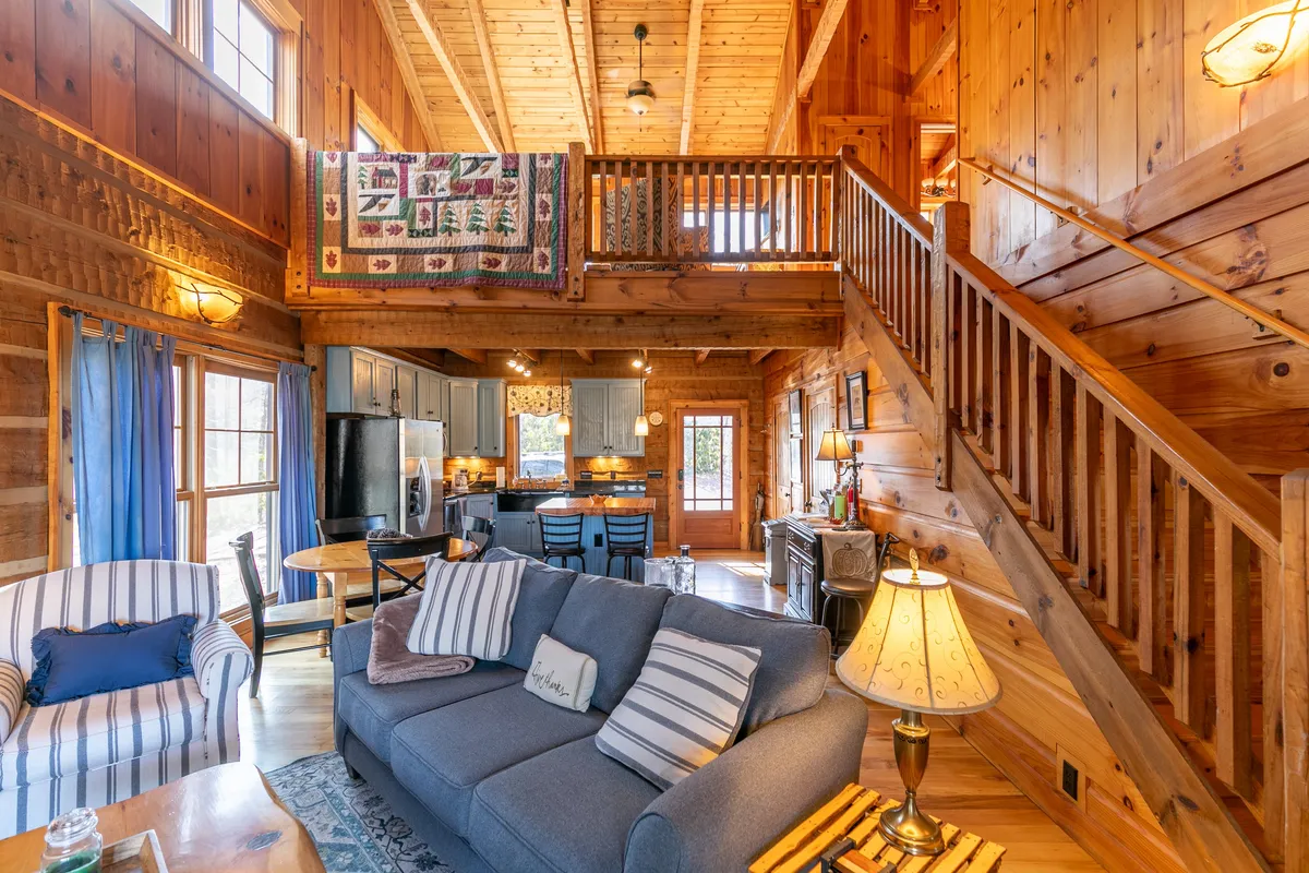 A warm, welcoming perspective of the living and dining area, complete with a comfortable sofa, a group dining table, and a glimpse of the kitchen and staircase. A decorative quilt adds a cozy touch, and the space feels perfect for shared meals, storytelling, and family time.