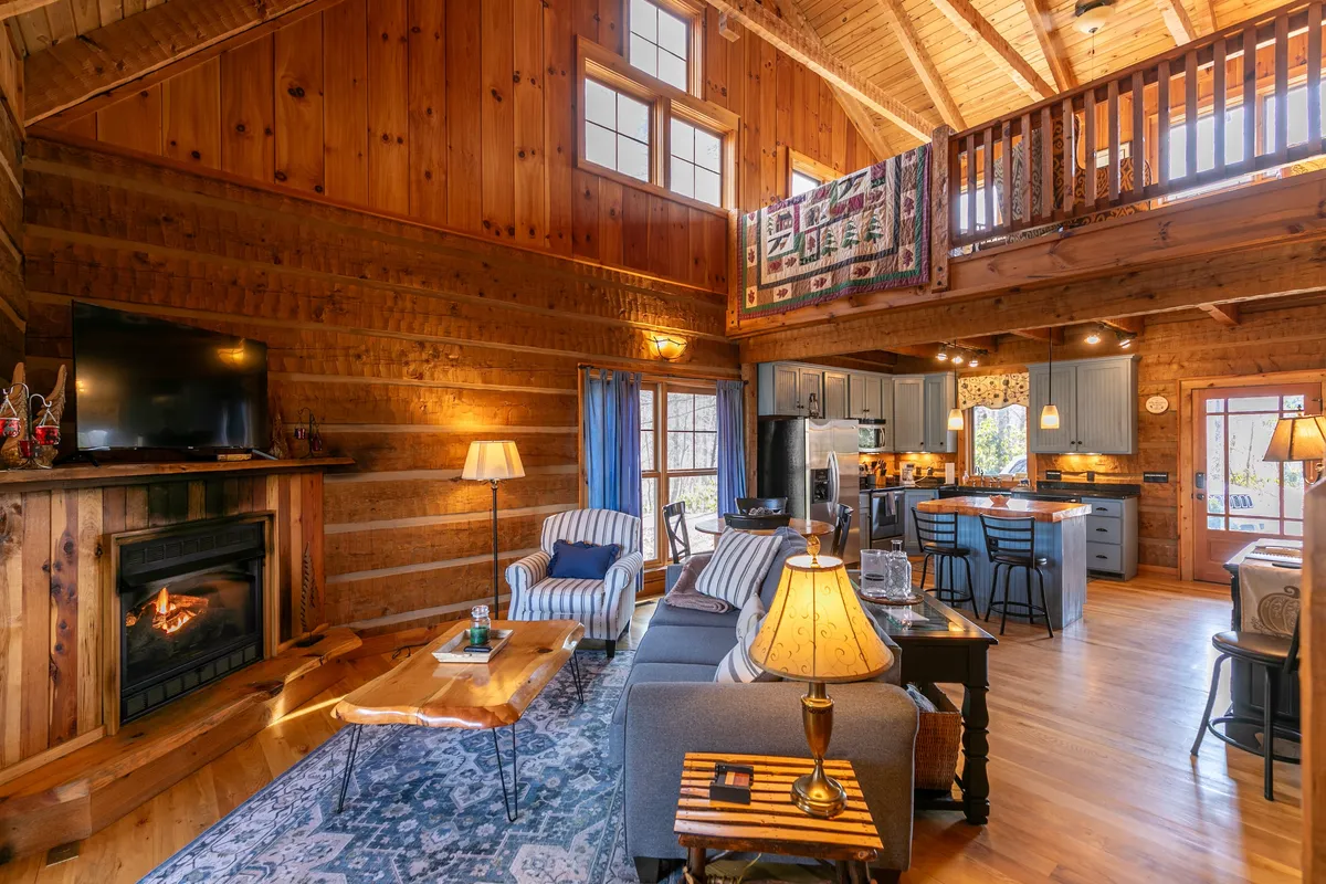 An inviting open-concept living area with a cozy gas fireplace, plush sofa, large windows that fill the space with natural light, and a view into the modern kitchen with a custom island. There is a quilt hanging from the balcony, adding a touch of homely charm. The scene exudes warmth and is perfect for gatherings, relaxation, and conversation.