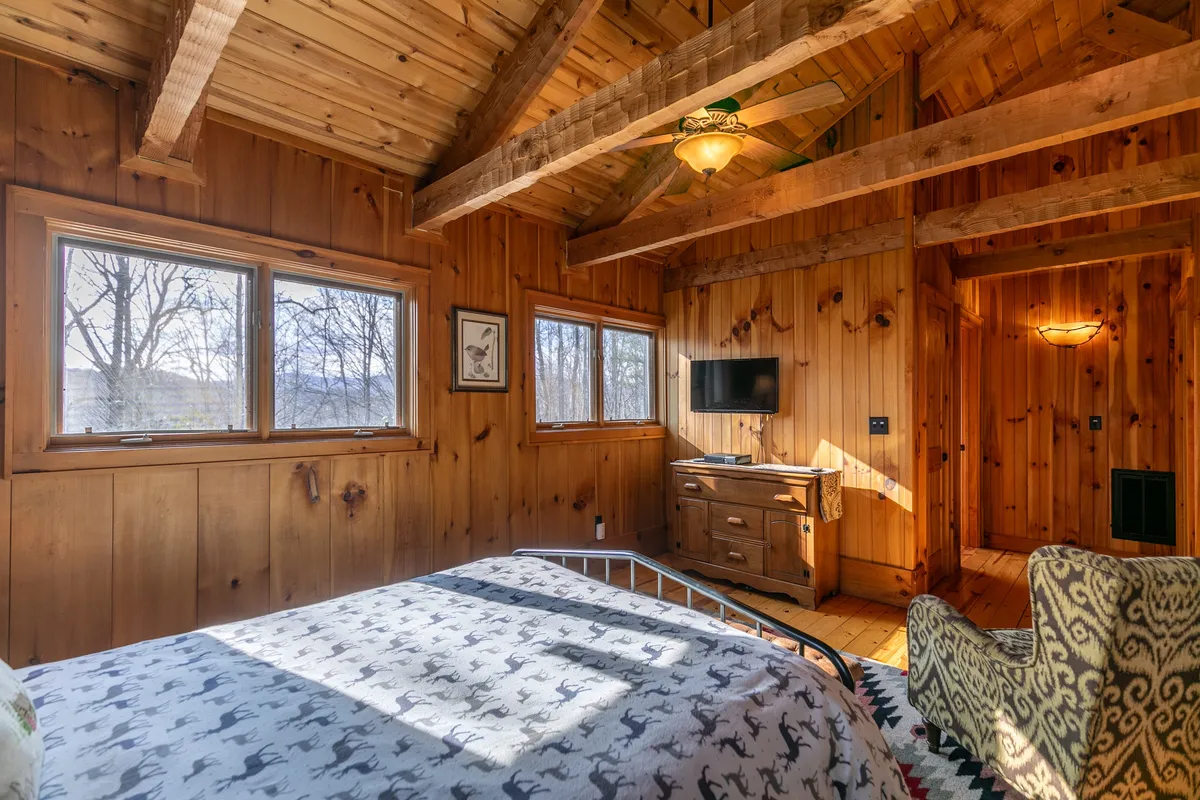 A cozy bedroom with a bed adorned in patterned bedding, sturdy wooden furniture, exposed ceiling beams, a wall-mounted TV, and generous natural light streaming through large windows. The warm, rustic decor is complemented by woodland views beyond the glass.