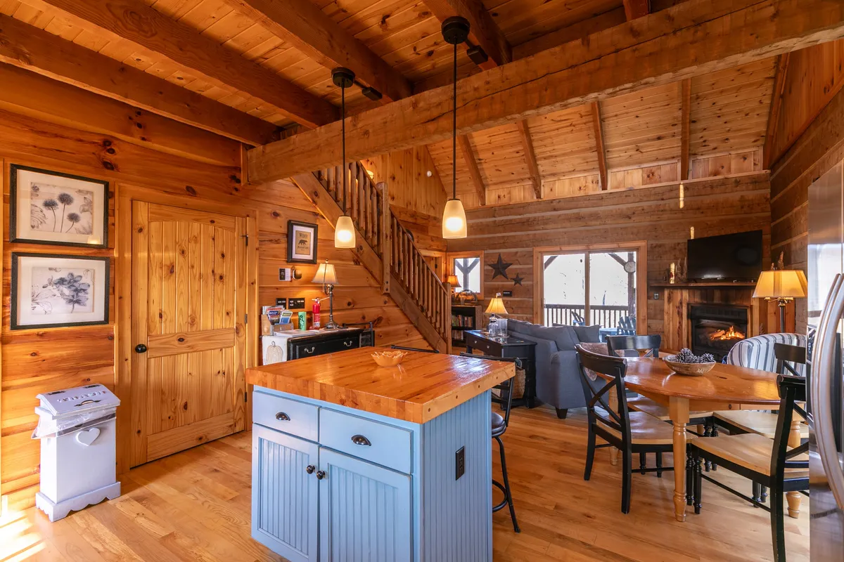 A welcoming open-concept space that includes a kitchen island, staircase, dining table, and fireplace. The arrangement promotes group interaction, with ample space for cooking, dining, and lounging. Large windows and access to the deck hint at the natural surroundings outside, linking the interior with the outdoors.