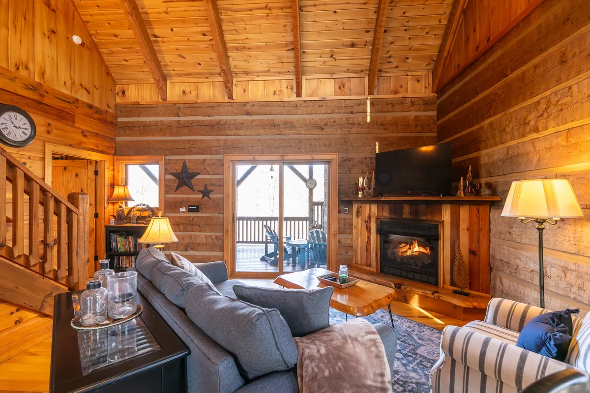 A warm, inviting living area with a crackling fireplace as the centerpiece, flanked by a cozy sofa, large windows revealing glimpses of the outdoors, wooden beams overhead, and charming decorative touches like a large clock and a star. The setting is perfect for unwinding together after a day of exploration, with the option to watch the sunset through the expansive windows or share quiet moments by the fire.