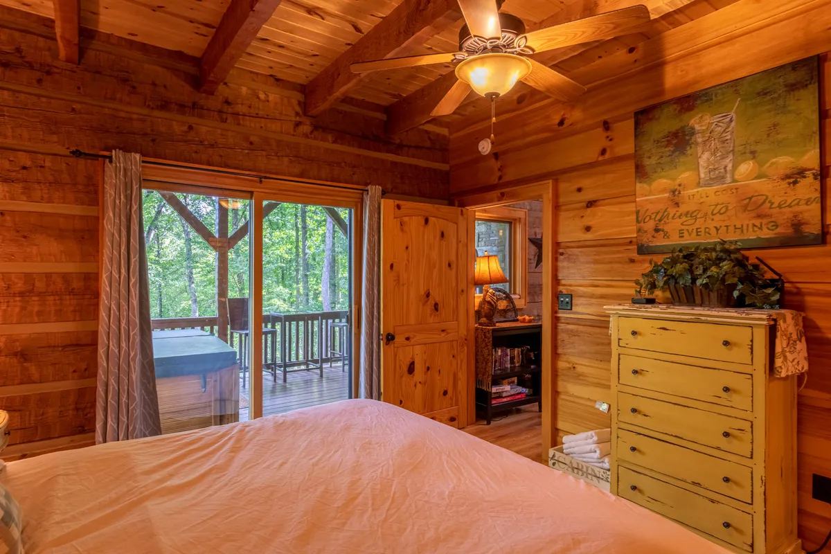 This interior image highlights a large window overlooking a lush natural landscape, wooden ceiling beams, a yellow dresser, and decorative artwork. The focus on the view through the window establishes a strong connection between the cozy interior and the outdoors.