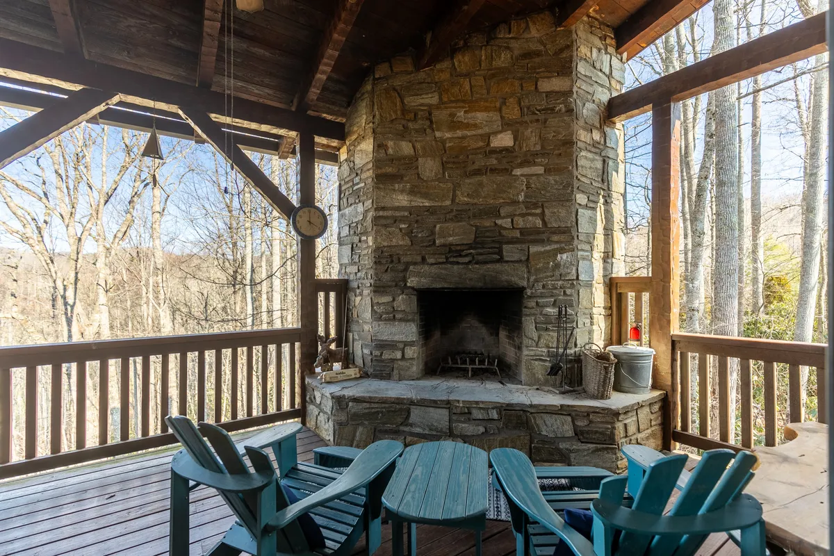 This image captures a cozy outdoor living space featuring a robust stone wood-burning fireplace, rustic Adirondack chairs, and tranquil woodland views. The deck is embraced by trees, creating a private and serene atmosphere that invites relaxation and gatherings around the fire.