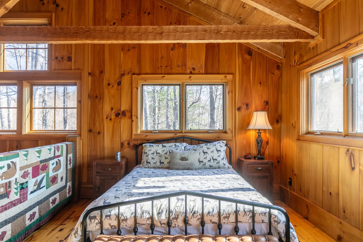 A bedroom with large windows framed by wooden walls, featuring a comfortable bed with decorative bedding and a quilted wall hanging. Natural light streams in, hinting at the changing seasons outside and inviting guests to relax while staying connected to the natural beauty beyond the windows.